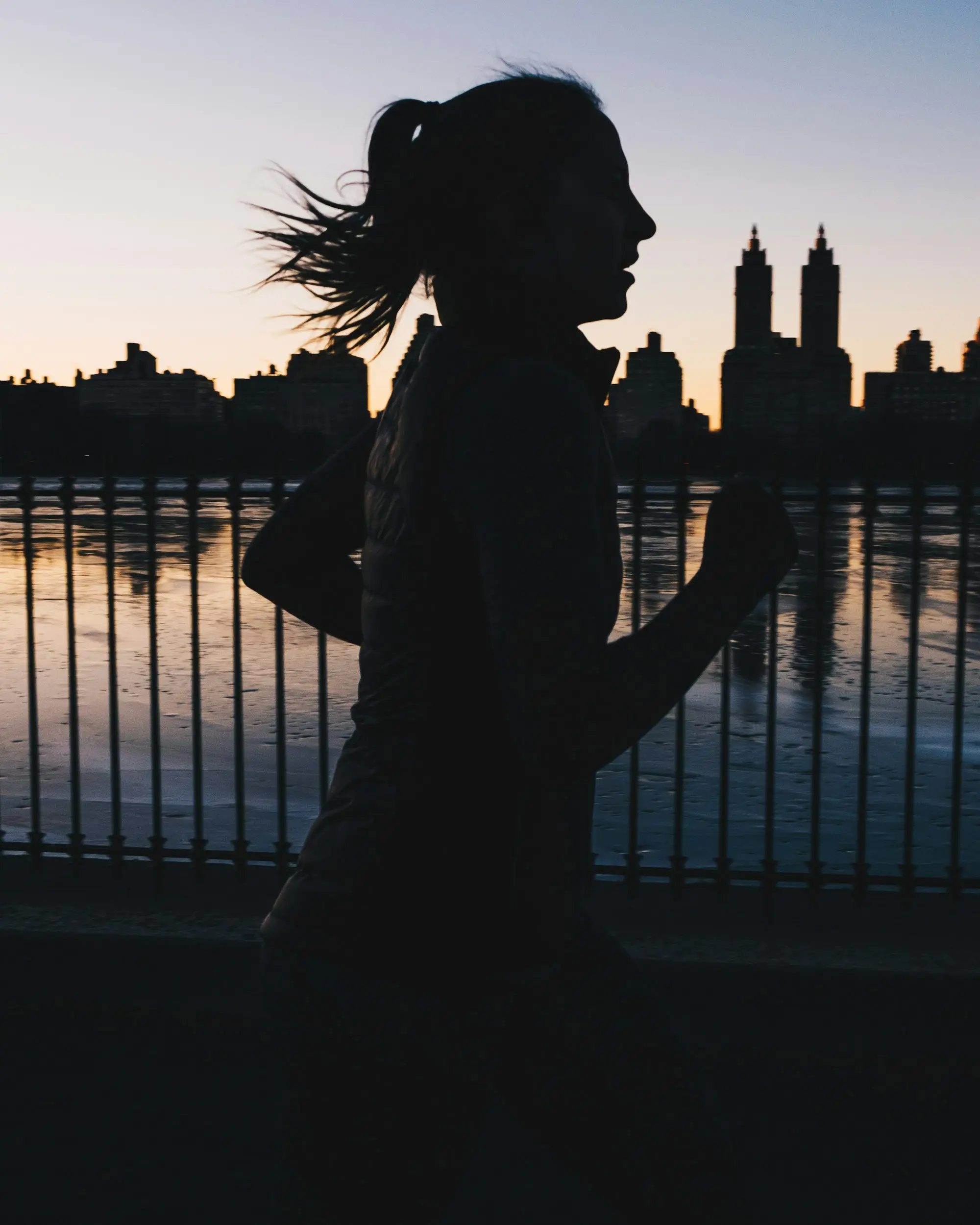 A runner in silhouette along a river at dusk, the skyline behind her